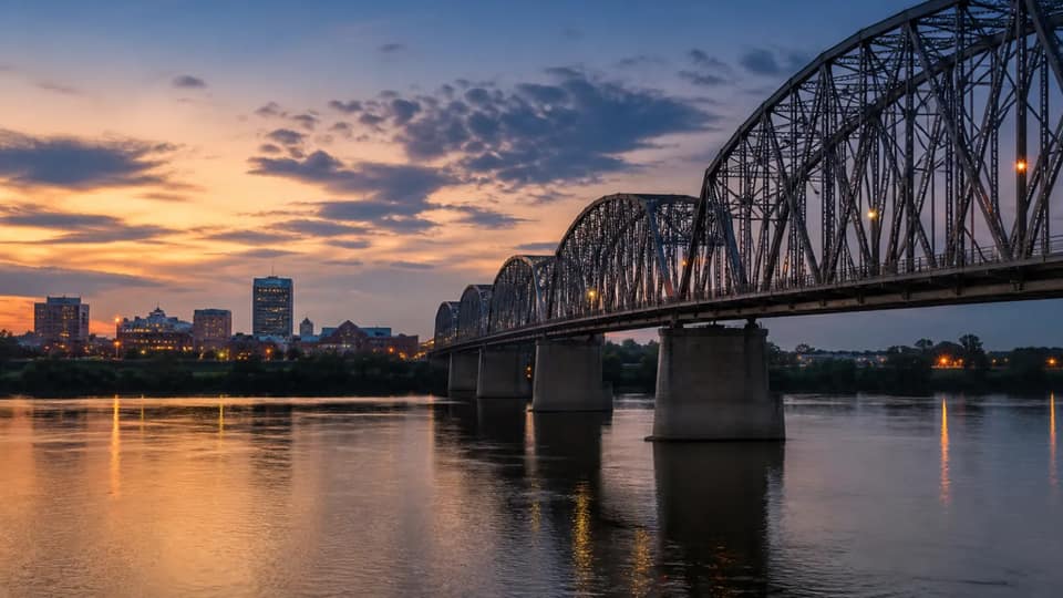 A steel truss bridge over a river at dusk, with a small city skyline lit up in the background