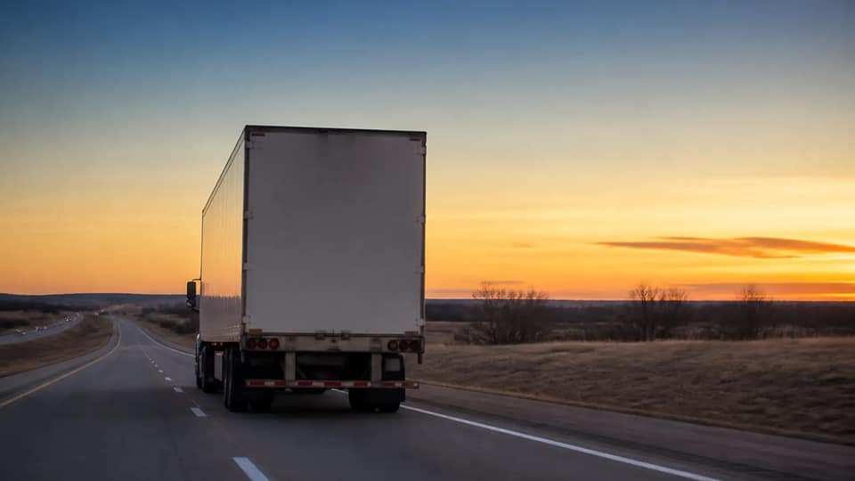 A large white truck driving on a two-lane highway across open plains at dusk
